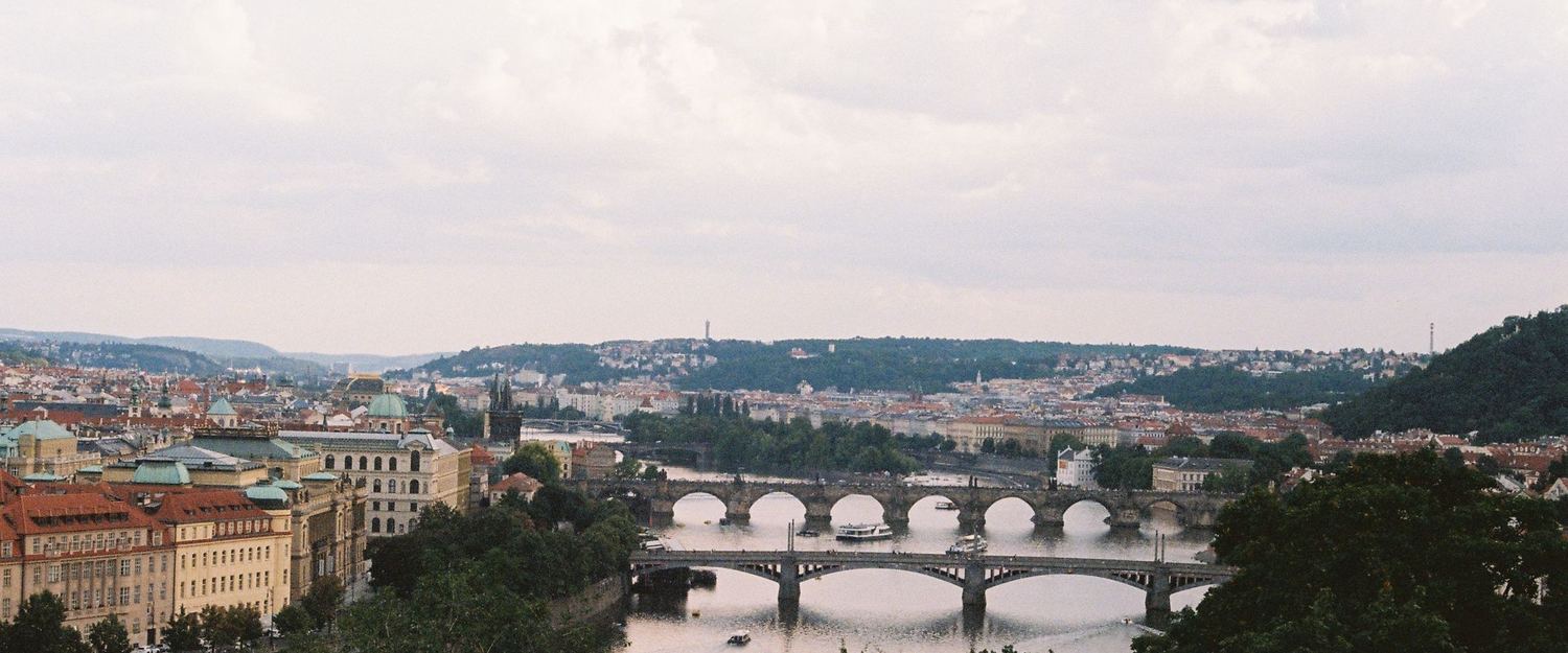 A European city seen from above its river: a stone bridge crossing still water, terracotta rooftops and pale facades receding along both banks under flat overcast light.